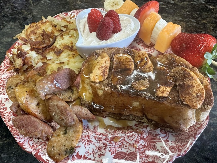 A delicious breakfast from Annville Inn, Bed and Breakfast. In this image, a red English china plate is loaded with baked French toast with Rosalie's carmel sauce, sweet potato hash, Italian sausage, fresh fruit skewer,and a heart of Cabot Yougurt from Vermont with three colossal raspberries on top.!