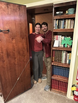 Couple standing in doorway of Secret Room, a hidden door behind a bookcase. Just engaged showing off engagement ring.