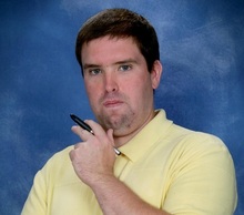 A head shot photo of Brian J Liston holding a pen in a thinking pose and wearing a yellow polo shirt.