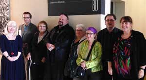 Image: photograph of eight people standing facing the camera and smiling. They stand insider under a sign which says Federal Court of Australia.
